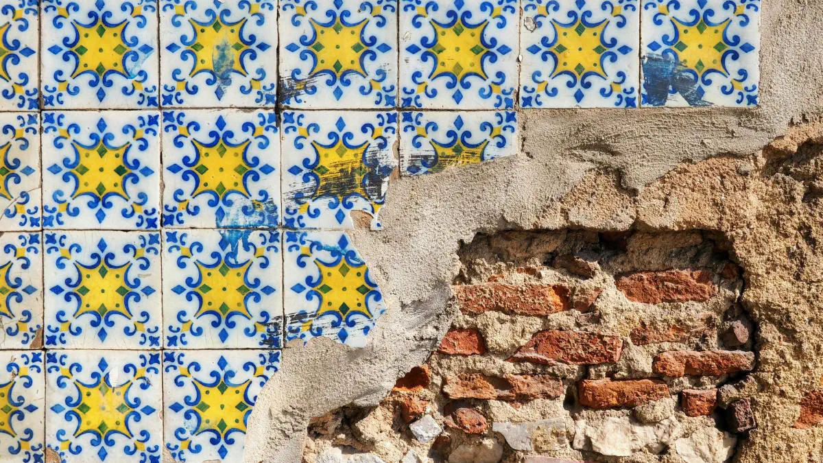 Close-up of a weathered wall where colorful blue-and-yellow patterned ceramic tiles are cracked and peeling away, revealing rough plaster and red brick underneath.