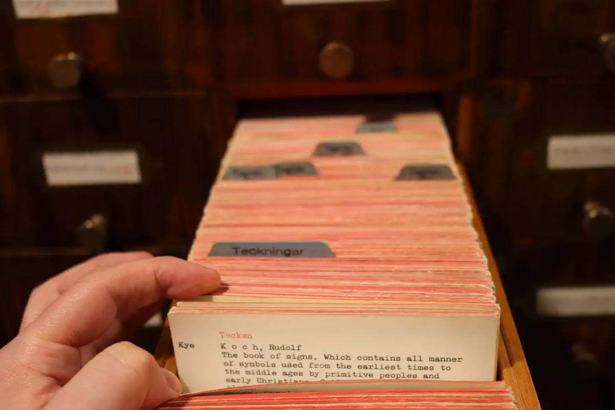 A hand pulling a labeled index card from the wooden drawer of an old library card catalog, with rows of cards fanned out behind it.